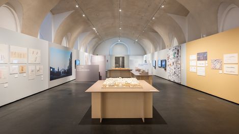 Exhibition room with high vaulted ceilings, multiple display panels on the walls, and a model on a table in the center