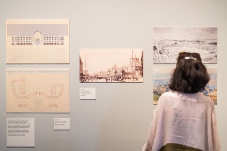 Person with dark hair and light pink top looking at several historical architectural drawings and city views on a light gray wall.