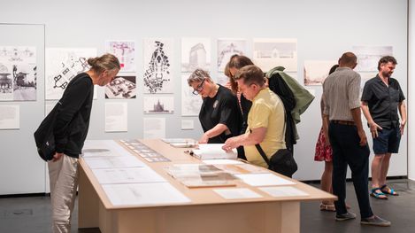 Several people examine drawings and documents spread out on a table in a gallery or exhibition with more images displayed on the wall behind them