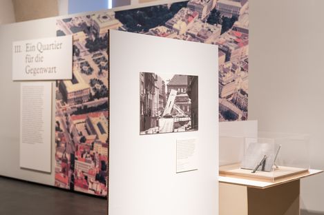 Exhibition with a photograph on a white panel, a large aerial city view in the background, and a sign reading 'III. Ein Quartier für die Gegenwart'
