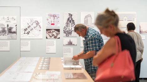 Several people examine plans and texts displayed on a white wall in an exhibition space.
