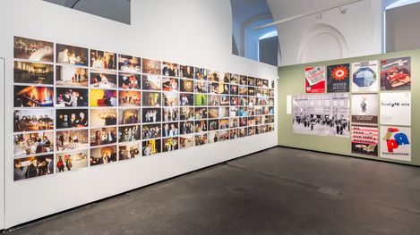 Gallery wall with numerous photos arranged in rows and adjacent wall with posters and texts in an exhibition space