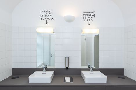 Zwei Waschbecken mit Spiegeln in einem weißen Badezimmer, über den Spiegeln steht jeweils handgeschriebener Text mit Pfeilen nach unten: 'IMAGINE YOURSELF 25 YEARS YOUNGER' links und 'IMAGINE YOURSELF 25 YEARS OLDER' rechts.