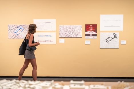 Frau in kurzem Kleid und Rucksack betrachtet Architekturzeichnungen an einer hellen Wand in einer Ausstellung