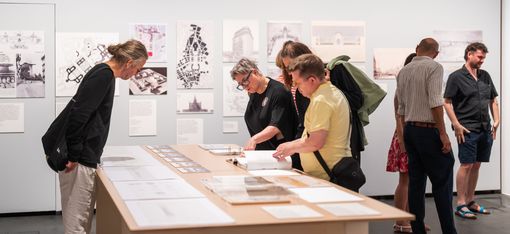 Several people examine drawings and documents spread out on a table in a gallery or exhibition with more images displayed on the wall behind them