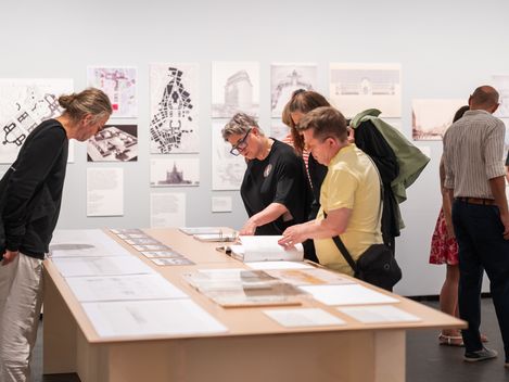 Several people examine drawings and documents spread out on a table in a gallery or exhibition with more images displayed on the wall behind them