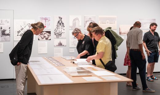 Mehrere Personen betrachten auf einem Tisch in einer Galerie oder Ausstellung ausgebreitete Zeichnungen und Dokumente, im Hintergrund hängen weitere Bilder an der Wand