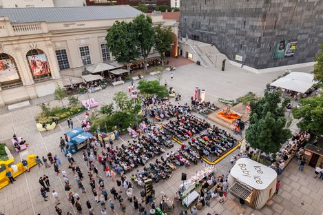 Bird's-eye view of a busy square with a stage hosting an event, surrounded by rows of seats and multiple people standing or sitting.
