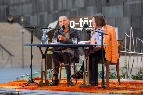 Two people sitting at a table with microphones speaking, a sign with 'o-töne' visible in the background