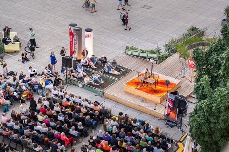 Stage with musician playing guitar in front of seated audience on a plaza with trees and plants