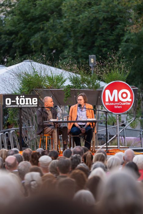 Two people sitting on a stage with microphones speaking to an outdoor audience with a red round sign reading 'MQ MuseumsQuartier Wien' in the background