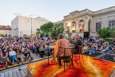 Two persons sit on chairs on an orange-red carpet facing a large outdoor audience in front of a historic building