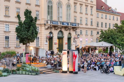 Large crowd seated in front of a historic building at an outdoor event with stage and lighting
