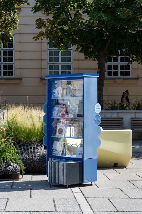 Blue public bookcase with glass front on a paved square in front of a building with large windows and trees in the background