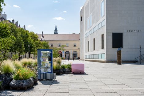 Public bookcase on a paved plaza area with benches and planted beds in front of a modern building labeled 'LEOPOLD'.
