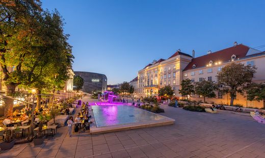 Abendlicher Haupthof des MuseumsQuartier Wien mit Wasserbecken, Gastronomie, Begrünung und beleuchteten Gebäuden.