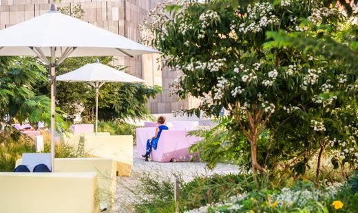 Person sitzt auf einer Bank in einem Garten mit Sonnenschirmen und blühenden Büschen vor einem modernen Gebäude