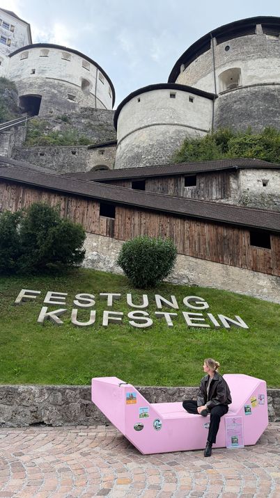 Eine Frau sitzt auf dem rosa MQ-Möbel vor der historischen Festung Kufstein. Auf dem grünen Hang dahinter steht der Schriftzug „Festung Kufstein“.