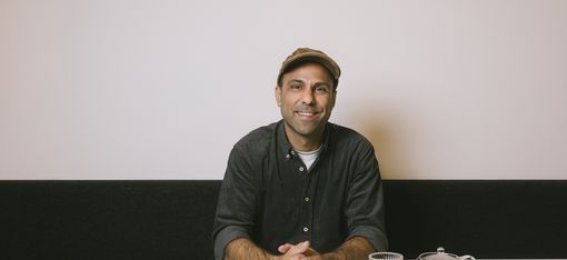Man wearing a flat cap sitting at a white table with a teapot and glass in front of a light-colored wall.