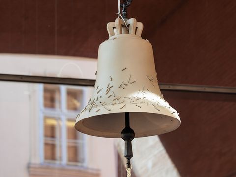 Large light beige bell with small irregularly scattered metal pins hanging in front of a window with white frames.
