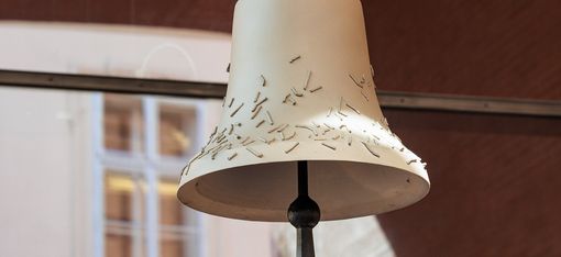 Large light beige bell with small irregularly scattered metal pins hanging in front of a window with white frames.