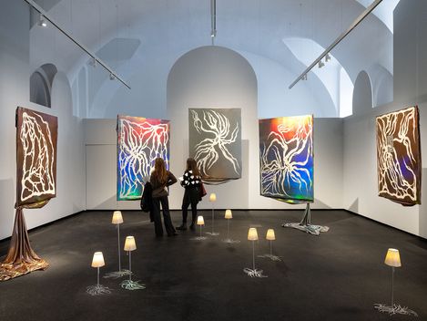 Zwei Personen betrachten in einem Ausstellungsraum fünf großformatige abstrakte Gemälde mit leuchtenden Linien auf dunklem Hintergrund, umgeben von kleinen Lampen auf dem Boden.