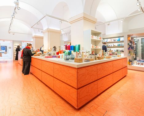 Museums­shop mit zentralem orangefarbenem Verkaufstresen und Besucherinnen, die Designprodukte und Bücher ansehen.