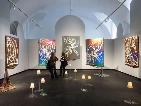 Two people observe five large abstract paintings with glowing lines on dark backgrounds in an exhibition room surrounded by small floor lamps.