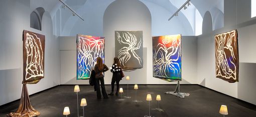 Zwei Personen betrachten in einem Ausstellungsraum fünf großformatige abstrakte Gemälde mit leuchtenden Linien auf dunklem Hintergrund, umgeben von kleinen Lampen auf dem Boden.