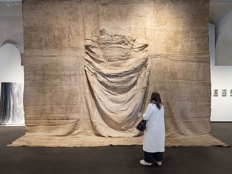Person in white coat stands in front of large textile-like artwork with folds in a gallery
