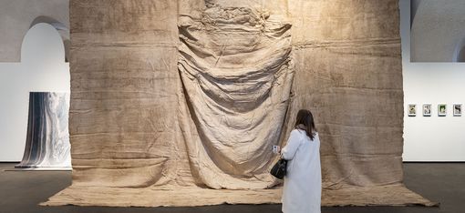 Person in weißem Mantel steht vor großem, textilähnlichem Kunstwerk mit Faltenwurf in einer Galerie