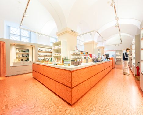 Großer Verkaufsbereich des MQ Point Shops mit zentralem Tresen und Besucher, der Produkte in den Regalen betrachtet.