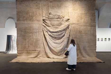 Person in weißem Mantel steht vor großem, textilähnlichem Kunstwerk mit Faltenwurf in einer Galerie