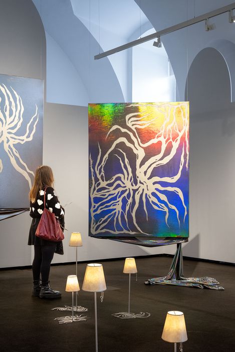 Frau mit Herzmuster-Pullover betrachtet in einem hellen Ausstellungsraum ein großes, schillerndes Kunstwerk mit wurzelartigen weißen Linien, umgeben von kleinen Lampen auf Ständern.
