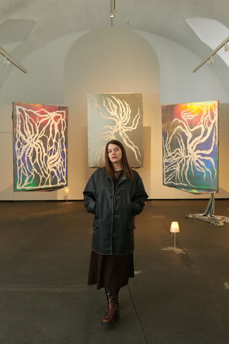 Person wearing a long coat and boots standing in an exhibition space with three abstract paintings on the wall and small lamps on the floor