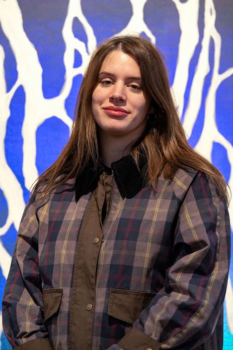 Portrait von Milica Živković. Person mit langen braunen Haaren trägt eine karierte Jacke mit braunen Taschen vor einem blau-weißen abstrakten Hintergrund