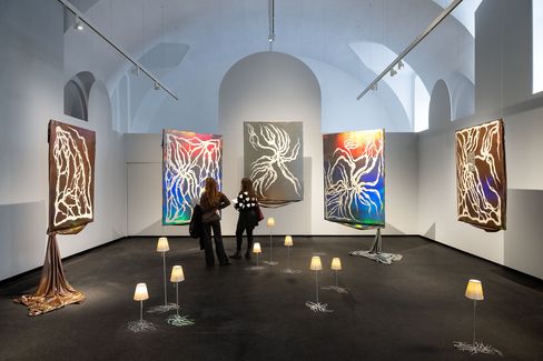Two people observe five large abstract paintings with glowing lines on dark backgrounds in an exhibition room surrounded by small floor lamps.