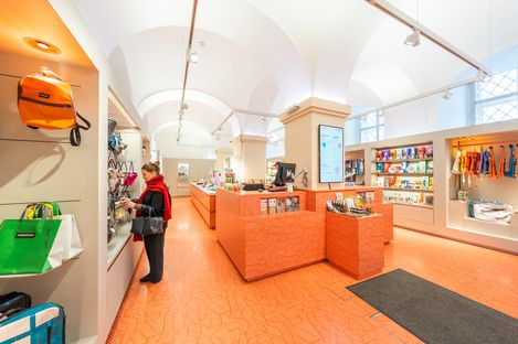 Innenansicht des MQ Point Shops mit orangefarbenem Verkaufstresen, hohen Gewölbedecken und Besucherin, die Taschen betrachtet.