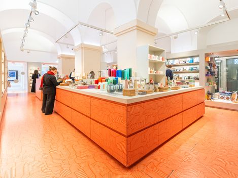 Museums­shop mit zentralem orangefarbenem Verkaufstresen und Besucherinnen, die Designprodukte und Bücher ansehen.