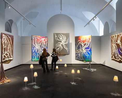 Two people observe five large abstract paintings with glowing lines on dark backgrounds in an exhibition room surrounded by small floor lamps.