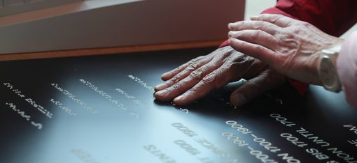 Zwei Hände berühren eine schwarze Tafel mit weißer Schrift, im Hintergrund ein Fenster.