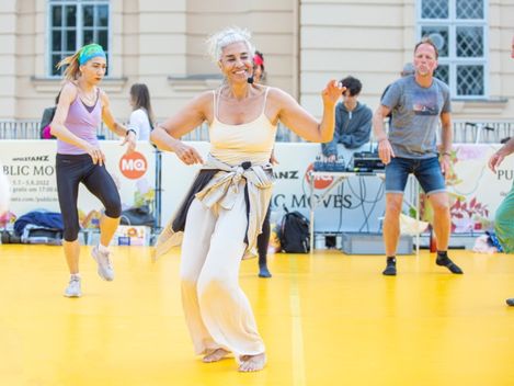 Several people dancing on a yellow floor in front of a white wall with windows and a DJ booth.