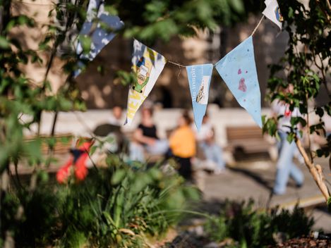 Nahaufnahme von bunten Wimpeln, die an einer Schnur hängen, unscharfer Hintergrund mit Menschen und Bänken in einem Park oder Garten
