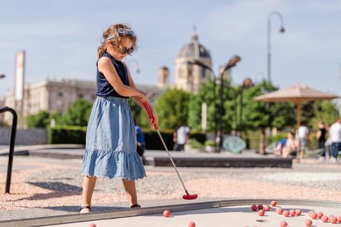 Child with curly hair and sleeveless top plays mini golf with a red club on an outdoor course