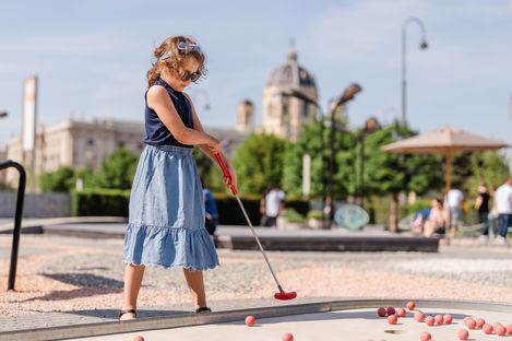 Child with curly hair and sleeveless top plays mini golf with a red club on an outdoor course