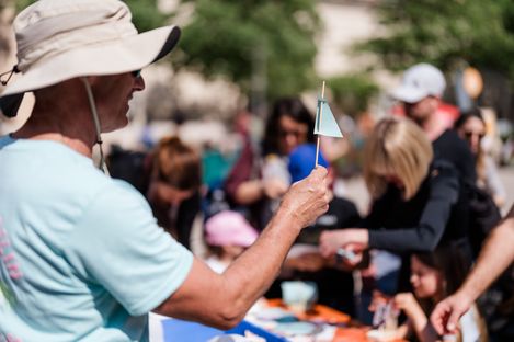 A person holds a small handmade paper sail while children work at a crafting table in the background.