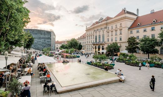 Belebter Haupthof des MuseumsQuartier bei Sonnenuntergang mit einem Spiegelbecken und Besuchern, die in der Nähe sitzen. Historische Gebäude und Bäume tragen zur ruhigen Atmosphäre bei.