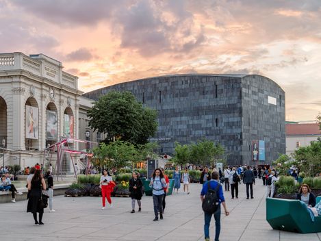 Menschen gehen und verweilen im belebten MQ Haupthof mit Bäumen und moderner sowie historischer Architektur bei Sonnenuntergang