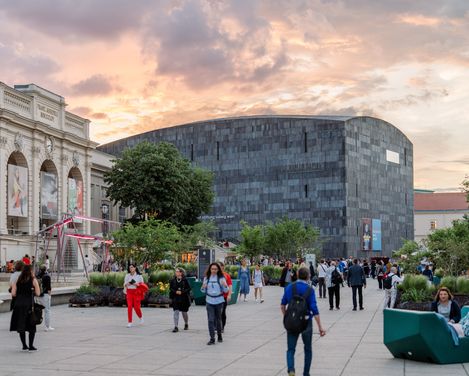 Menschen gehen und verweilen im belebten MQ Haupthof mit Bäumen und moderner sowie historischer Architektur bei Sonnenuntergang