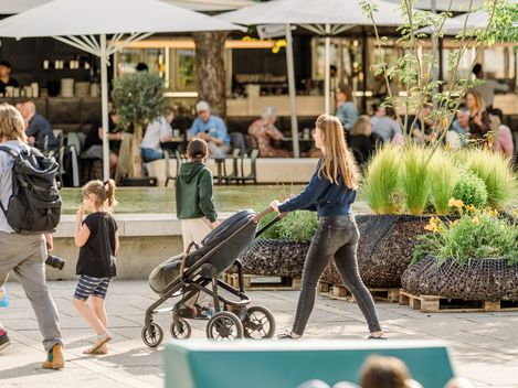 Frau mit Kinderwagen und Kind an der Hand geht durch belebten Außenbereich mit Sonnenschirmen und Sitzgruppen.